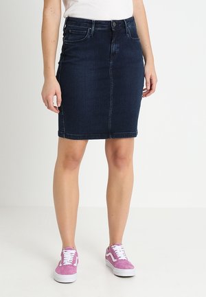 Femme portant une jupe en jean bleu foncé longueur genou et des baskets roses, debout sur un sol blanc avec un fond blanc.