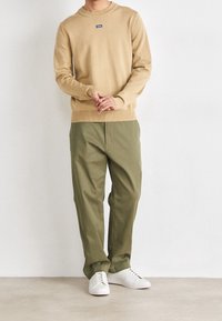 Beige villapaita, jossa on pyöreä kaula-aukko ja pieni sininen logo, yhdistettynä löysiin oliivinvihreisiin housuihin ja valkoisiin lenkkareihin.