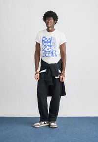 Tricou alb cu imprimeu albastru, pantaloni de trening negri și adidași Nike. Hanorac legat în jurul taliei; material texturat, croială lejeră.