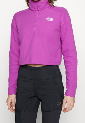 Mujer con un forro polar corto púrpura con cuello con cremallera y pantalones deportivos negros de cintura alta con bolsillos con cremallera.
