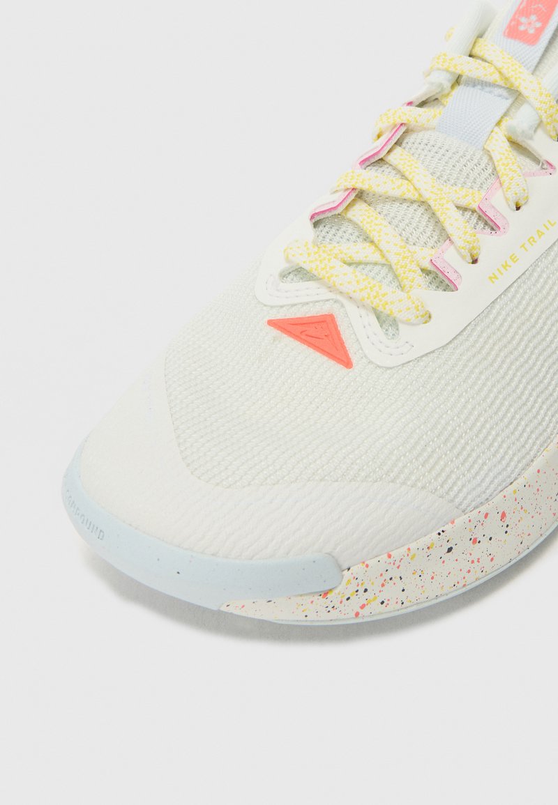 Zapatilla deportiva gris claro con parte superior texturizada, cordones amarillos y rosas, y una entresuela de espuma multicolor salpicada. Presenta un logo en el costado.