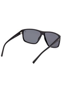 Gafas de sol rectangulares negras con lentes gris oscuro, brazos delgados y un acabado mate. Presentan un logotipo sutil en el brazo.