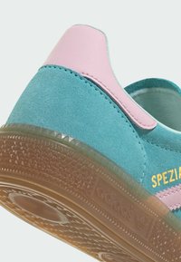 Gros plan sur une chaussure Adidas en daim bleu sarcelle avec languette de talon rose et semelle translucide marron, présentant le texte doré "SPEZIAL" sur le côté.