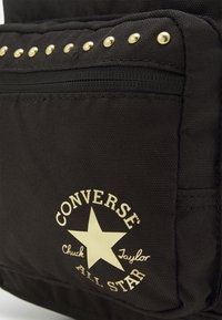 Sac à dos en tissu noir avec des clous dorés, poche zippée et logo blanc proéminent avec une étoile. Texture durable et design simple.