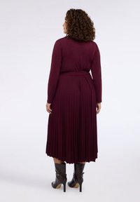 Bordowa plisowana sukienka z długimi rękawami, talią podkreśloną paskiem i gładką fakturą, zestawiona z czarnymi, sięgającymi do kolan botkami na obcasie.