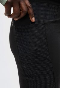 Χέρι με σκουρόχρωμο δέρμα που τοποθετεί ένα μαύρο smartphone στην μαύρη, πουα πλαϊνή τσέπη εφαρμοσμένων παντελονιών.