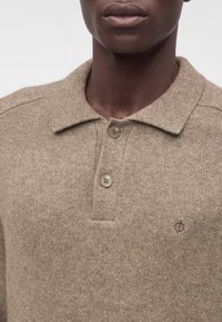 Personne à la peau foncée portant un pull polo en maille beige avec deux boutons et un petit logo brodé sur la poitrine.