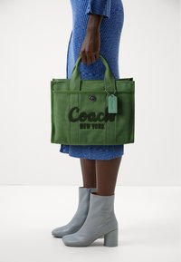 Grön tygkasse med broderad text "Coach New York". Har dubbla topplankar och en mintgrön etikett. Bärs med blå ribbstickad klänning och gråa ankelkängor.