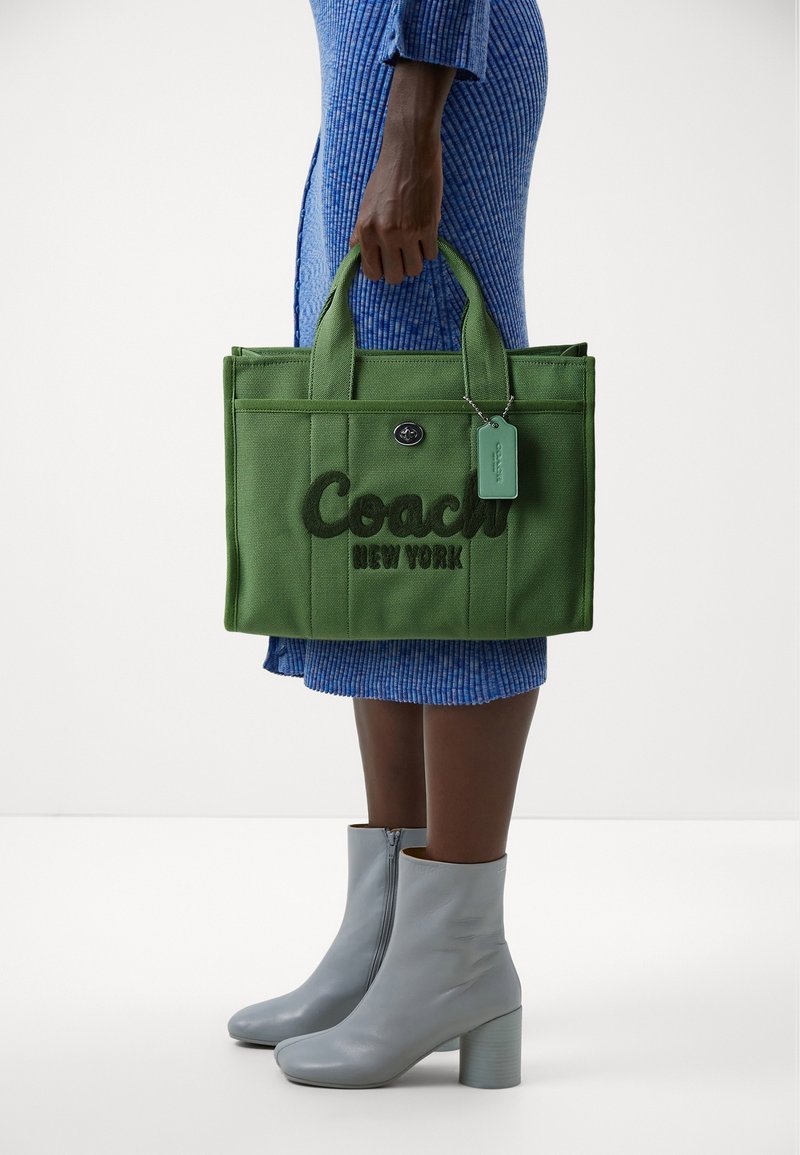 Grön tygkasse med broderad text "Coach New York". Har dubbla topplankar och en mintgrön etikett. Bärs med blå ribbstickad klänning och gråa ankelkängor.