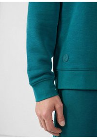 Teal sweatshirt met ribgebreide manchetten en zoom, met een klein, reliëflogo op de onderste zoom. Glad textuur, casual ontwerp.