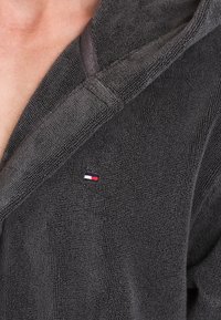 Grå frottébadekåbe med hætte, der har et tekstureret stof og et lille logo i marineblå, rød og hvid på venstre side.