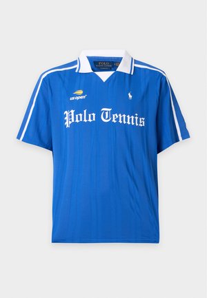 Homme debout portant un polo de tennis bleu, un pantalon bleu marine et des baskets blanches sur un fond blanc uni.