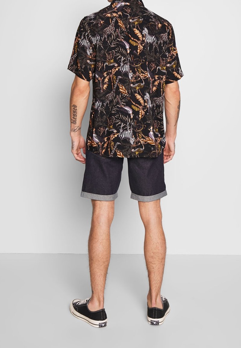 Camisa negra de manga corta con estampado colorido de animales y follaje, combinada con pantalones cortos de mezclilla oscuros con los dobladillos enrollados y zapatillas negras.