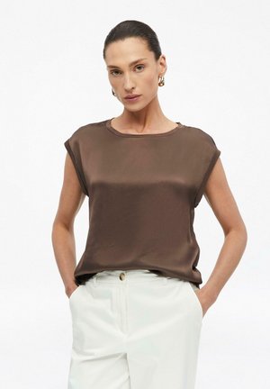 Femme aux cheveux plaqués en arrière, portant des boucles d'oreilles créoles en or, un chemisier marron sans manches et un pantalon blanc, les mains dans les poches.