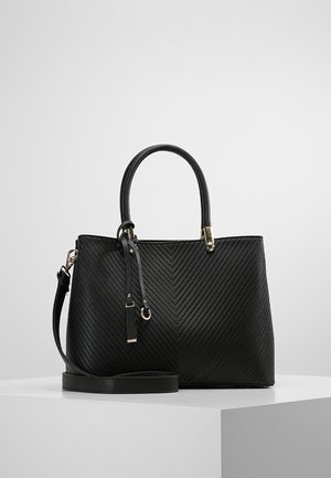 Sac à main en cuir noir avec motif de couture en chevron, deux poignées, quincaillerie dorée et bandoulière amovible sur surface d'exposition blanche.