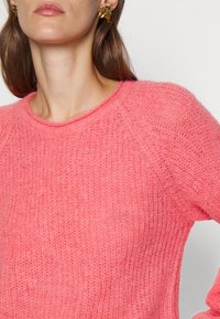 Frau in einem strukturierten pinken Strickpullover und goldenen geometrischen Ohrringen, von Hals bis zur Mitte des Oberkörpers vor einem weißen Hintergrund gezeigt.