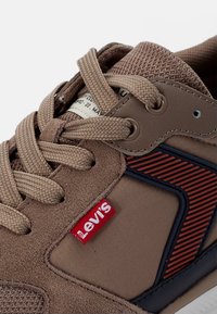 Brązowy sneaker z teksturowaną cholewką z zamszu, siateczkowym językiem i kontrastującymi granatowymi akcentami. Posiada beżowe sznurowadła oraz czerwoną metkę z logo Levi's.