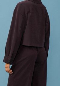 Person iført en mørk lilla langærmet cropped jakke og matchende vide bukser, stående mod en blå baggrund.