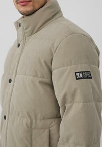 Beige Steppjacke mit Cord-Textur, hohem Kragen, schwarzen Knöpfen und einem Logo-Patch auf dem linken Ärmel.