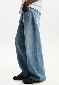 Light blue denim jeans with a wide-leg silhouette, featuring a classic five-pocket design and a faded texture. Brown footwear visible.