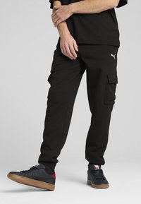 Schwarze Cargo-Hose aus weichem Stoff mit zwei Seitentaschen und einem Logo auf dem Oberschenkel. Kombiniert mit dunklen Turnschuhen mit Gummisohlen.