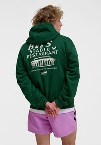 Green hoodie with white text and graphic on the back, paired with pink shorts. The hoodie features a kangaroo pocket and ribbed cuffs.