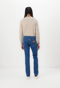 Tricou lung bej deschis, blugi din denim albastru cu o croială standard, cinci buzunare și o etichetă maro pe spate. Design curat, clasic.