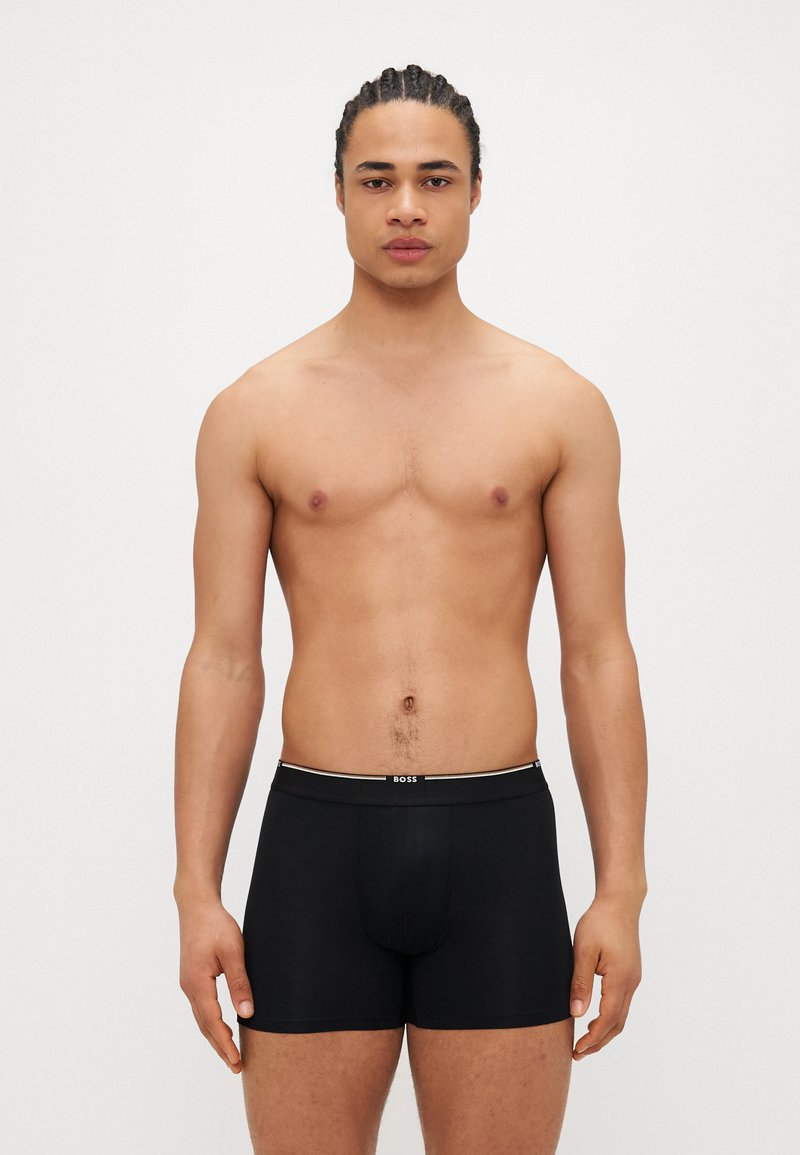 Schwarze Boxershorts mit einem Logo-Gummibund, die eine glatte Textur und eine figurbetonte Passform aufweisen, getragen von einem männlichen Modell.