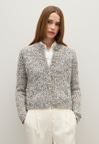 Cárdigan de punto gris y blanco con cuello redondo, decorado con grandes botones de madera y puños acanalados, llevado sobre una camisa blanca.