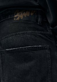 Musta farkkuhousut, joissa on kulunut taskunreuna, kuvioitu kankaan pinta ja suorakulmainen musta nahkalappu graafisella logolla.