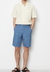 Mężczyzna w kremowej, krótko rękawowej koszuli z guzikami, niebieskich szortach do kolan i czarnych sandałach, stojący z rękami w kieszeniach.