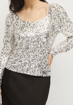 Frau trägt ein silbernes Pailletten-Oberteil mit langen Ärmeln und einem Rundhalsausschnitt sowie einen schlichten schwarzen Rock, die Hand auf der Hüfte ruhend.