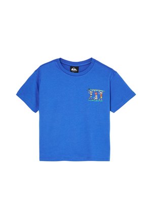 Młody chłopiec z kręconymi włosami, ubrany w niebieski t-shirt z grafiką, luźne ciemne dżinsy i czarne buty, stoi z rękami w kieszeniach na białym tle.