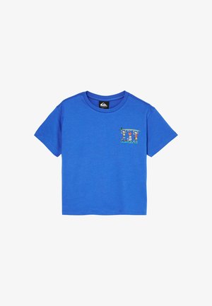 Młody chłopiec z kręconymi włosami, ubrany w niebieski t-shirt z grafiką, luźne ciemne dżinsy i czarne buty, stoi z rękami w kieszeniach na białym tle.