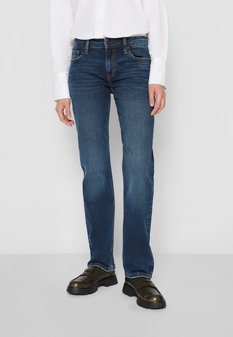 Jean en denim bleu foncé avec une coupe droite, cinq poches et des coutures contrastantes. Porté avec des chaussures noires sans lacets.
