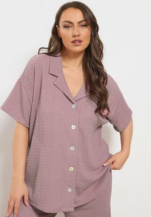 Frau mit langen braunen Haaren, die ein strukturiertes, mauvefarbenes kurzärmliges Hemd mit Knöpfen und passende Hosen trägt und vor einem schlichten, hellen Hintergrund steht.