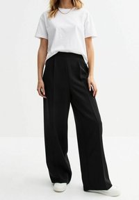 Schwarze weite Hose mit hoher Taille, aus glattem Stoff und mit gefaltetem Frontteil, kombiniert mit einem schlichten weißen T-Shirt und weißen Sneakern.