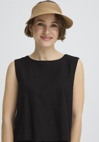 Femme aux cheveux bruns courts, portant un haut noir sans manches, des boucles d'oreilles en or et une visière tissée couleur bronzée, souriant doucement devant un fond blanc.
