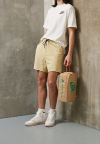 Beiga shorts med svart dragsko, vit Nike-t-shirt med liten logotyp, krämfärgade sneakers och en beige väska med grön branding.