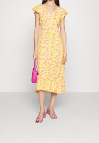 Robe midi florale jaune avec des manches courtes, ourlet à volants et décolleté en V, associée à un sac à main rose et des sandales à talons blocs transparentes.