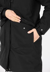 Svart jacka med dragkedja, två frampockets med tryckknappar och ett strukturerat tyg. Hand delvis synlig inne i en ficka.