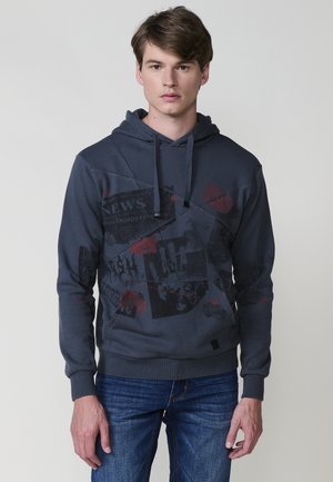 Sudadera gris oscuro con un bolsillo frontal, tejido texturizado y un estampado gráfico que presenta motivos de periódico y acentos rojos.
