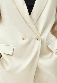 Krémově zbarvený dvouřadový blazer se dvěma průhlednými knoflíky, s klopami a bočními kapsami. Hladká, lehká tkanina s jemným leskem.