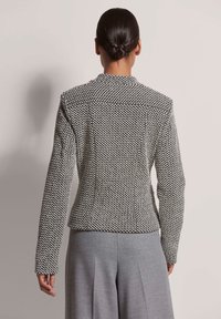 Schwarz-weiß karierter Blazer im Hahnentrittmuster mit taillierter Silhouette, Reverskragen und langen Ärmeln, kombiniert mit grauen Hosen.