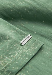 Vihreä kangas, jossa on hienovarainen hajautettu geometrinen kuosi. Sisältää hopeisen tarran, jossa lukee "ombre dept." kohokuvioituna, mikä korostaa sileää pintaa.