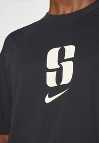 Juoda medvilninė marškinėliai su ryškiu, kreminės spalvos grafiniu elementu "S" ir mažuoju Nike swoosh logotipu priekyje. Trumpi rankovės, apvalus kaklas.