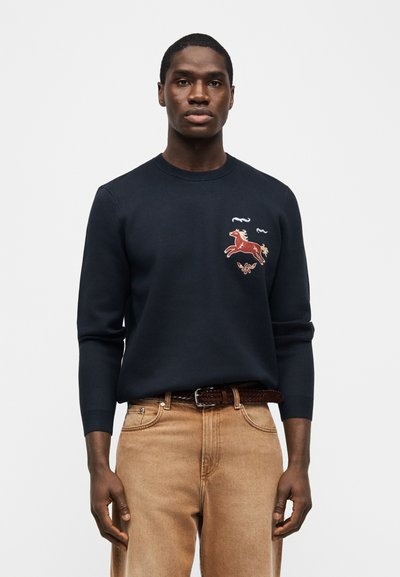 Άνδρας που φοράει μπλε πουλόβερ με κεντημένο κόκκινο άλογο και πεταλούδα, συνδυασμένο με τζιν ανοιχτό καφέ και πλεγτή δερμάτινη ζώνη, στέκεται μπροστά σε λευκό φόντο.