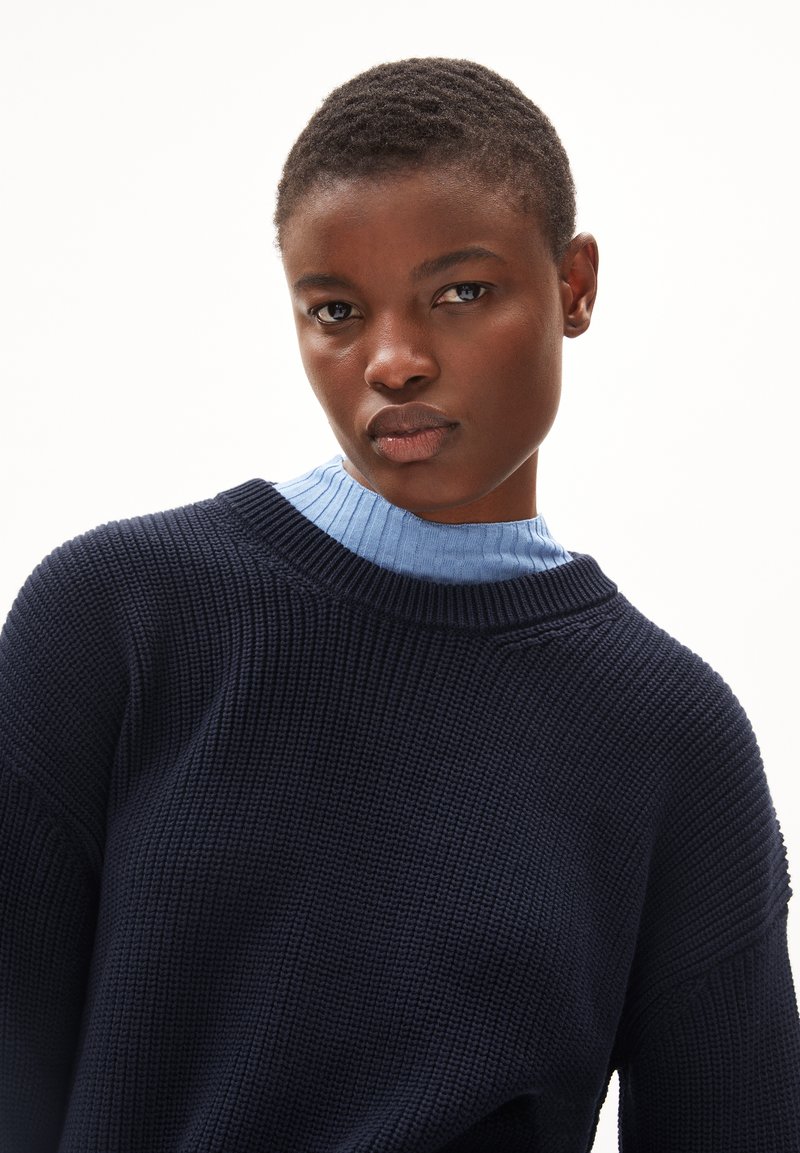 Marineblaue Strickjacke mit strukturiertem Muster, runder Ausschnitt und dezenten Rippakzenten am Kragen. Hellblaues Unterhemd sichtbar.