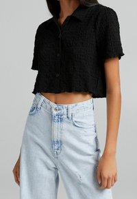 Donna che indossa una camicia nera a maniche corte, cropped e testurizzata, abbinata a jeans in denim azzurro chiaro a vita alta con dettagli sfilacciati.