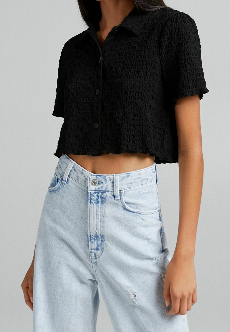 Donna che indossa una camicia nera a maniche corte, cropped e testurizzata, abbinata a jeans in denim azzurro chiaro a vita alta con dettagli sfilacciati.